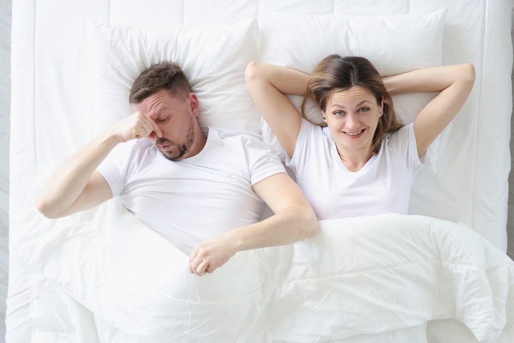 Married couple in bed, with the man covering his nose